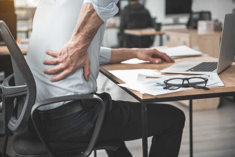 Gros plan de profil d'un homme assis à son bureau, une main crispée sur le bas du dos à cause d'une douleur lombaire
