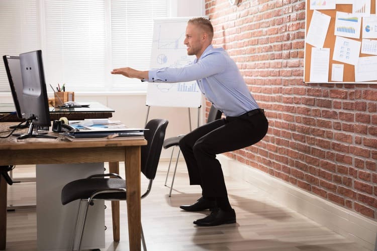 Un homme en chemise et pantalon de costume effectue un exercice de squat devant son bureau pendant une pause active au travail